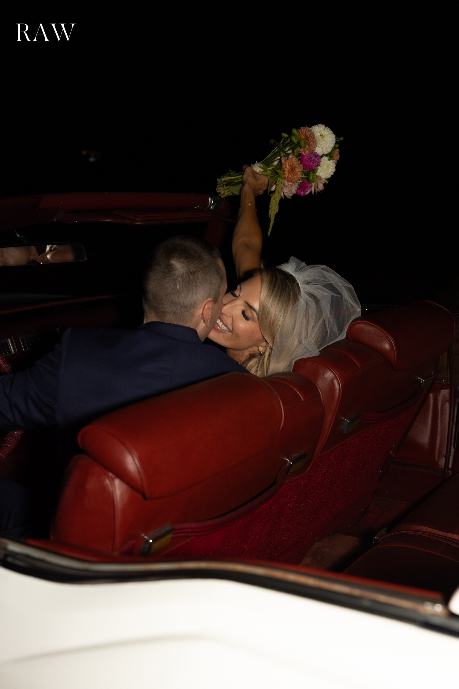 Wedding couple reclining in a vintage car with red leather seats at night.