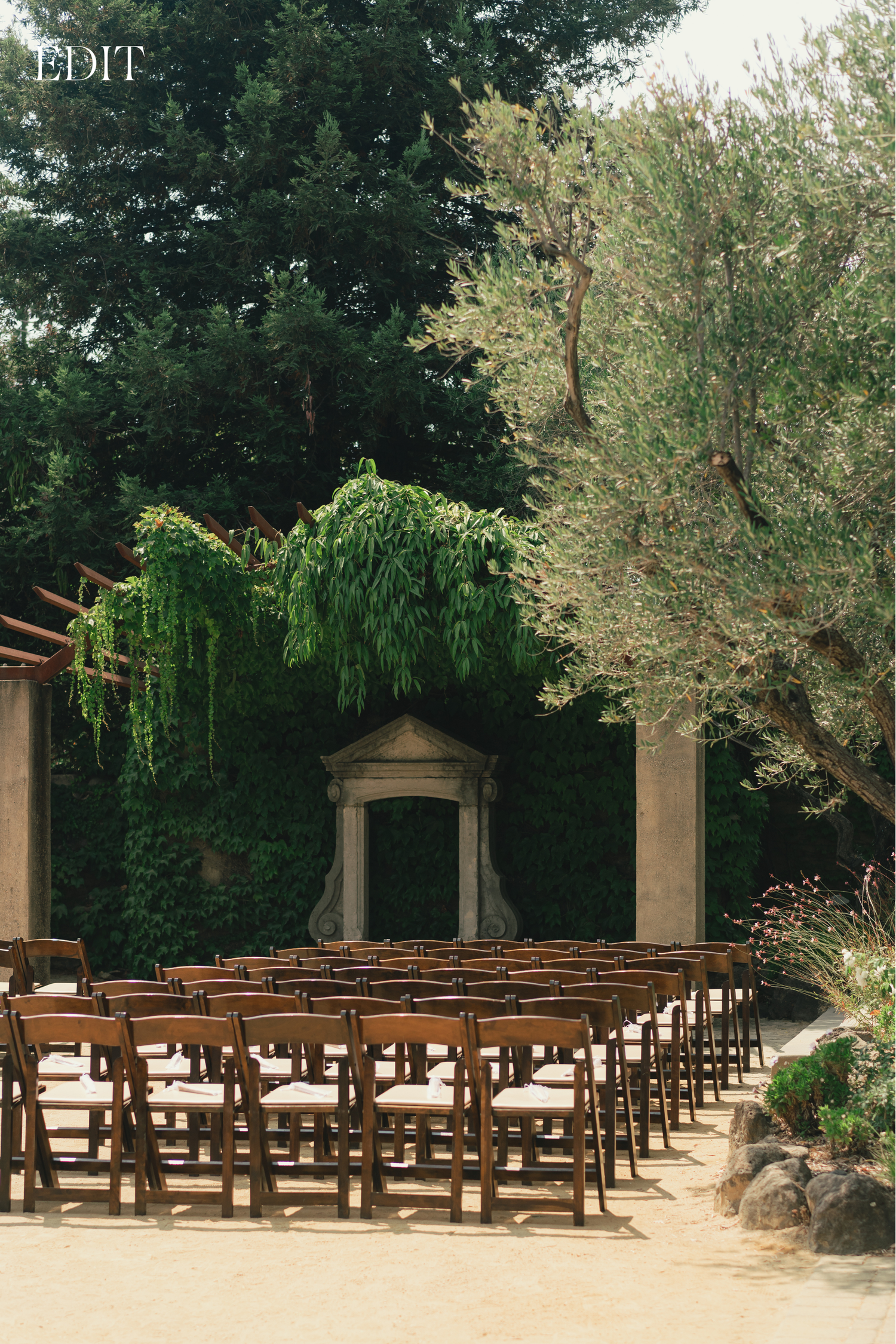 Wooden ceremony chairs arranged in rows beneath towering trees and stone pillars.