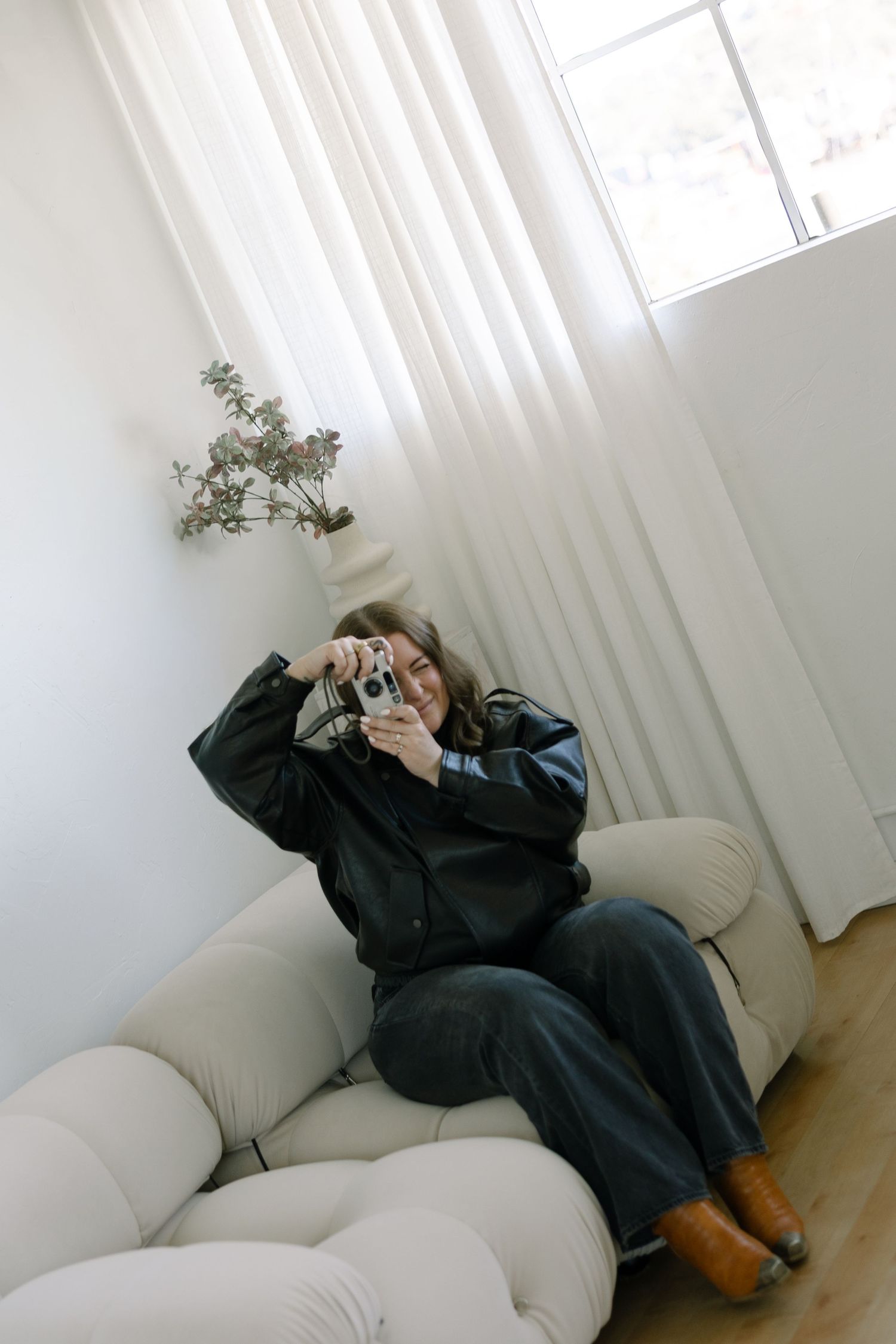 A person in dark clothing sits on a white modular sofa taking a photo with a vintage camera near a bright window.
