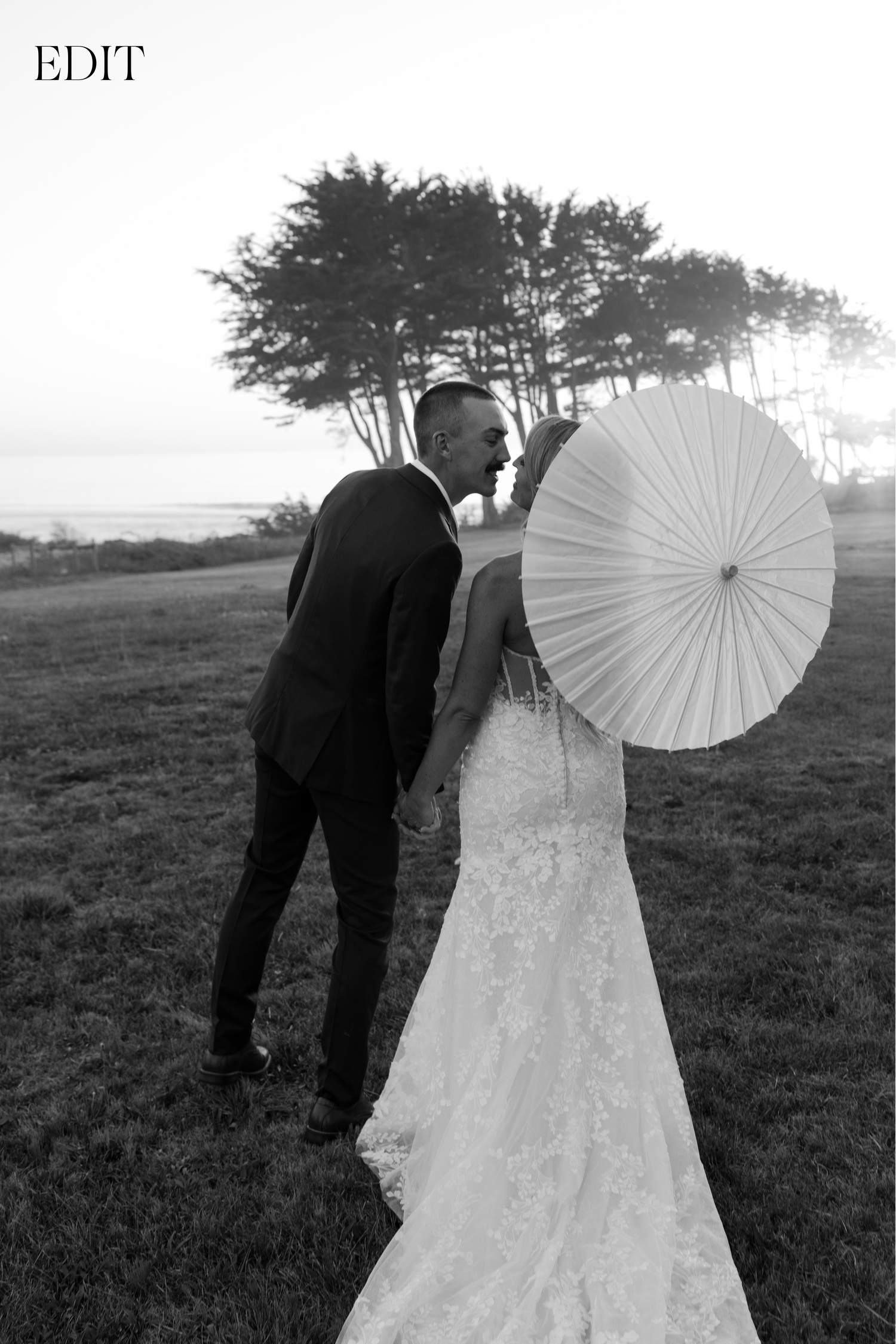 Black and white photo of newlyweds with a white parasol in an outdoor field setting.