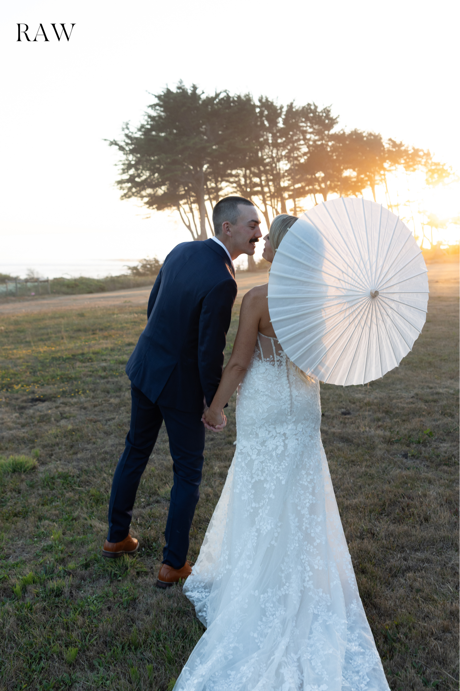 Couple in wedding attire walking through a field at sunset with a white parasol.
