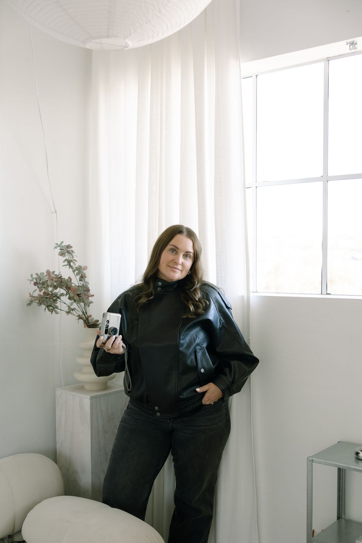 Person wearing black outfit poses in bright white room with sheer curtains and window light.