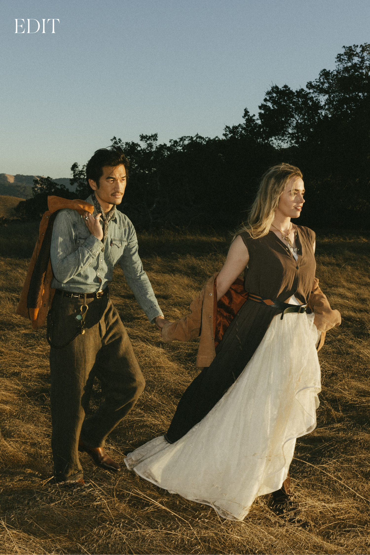Stylized photo of two people in vintage attire walking through a sunlit meadow.