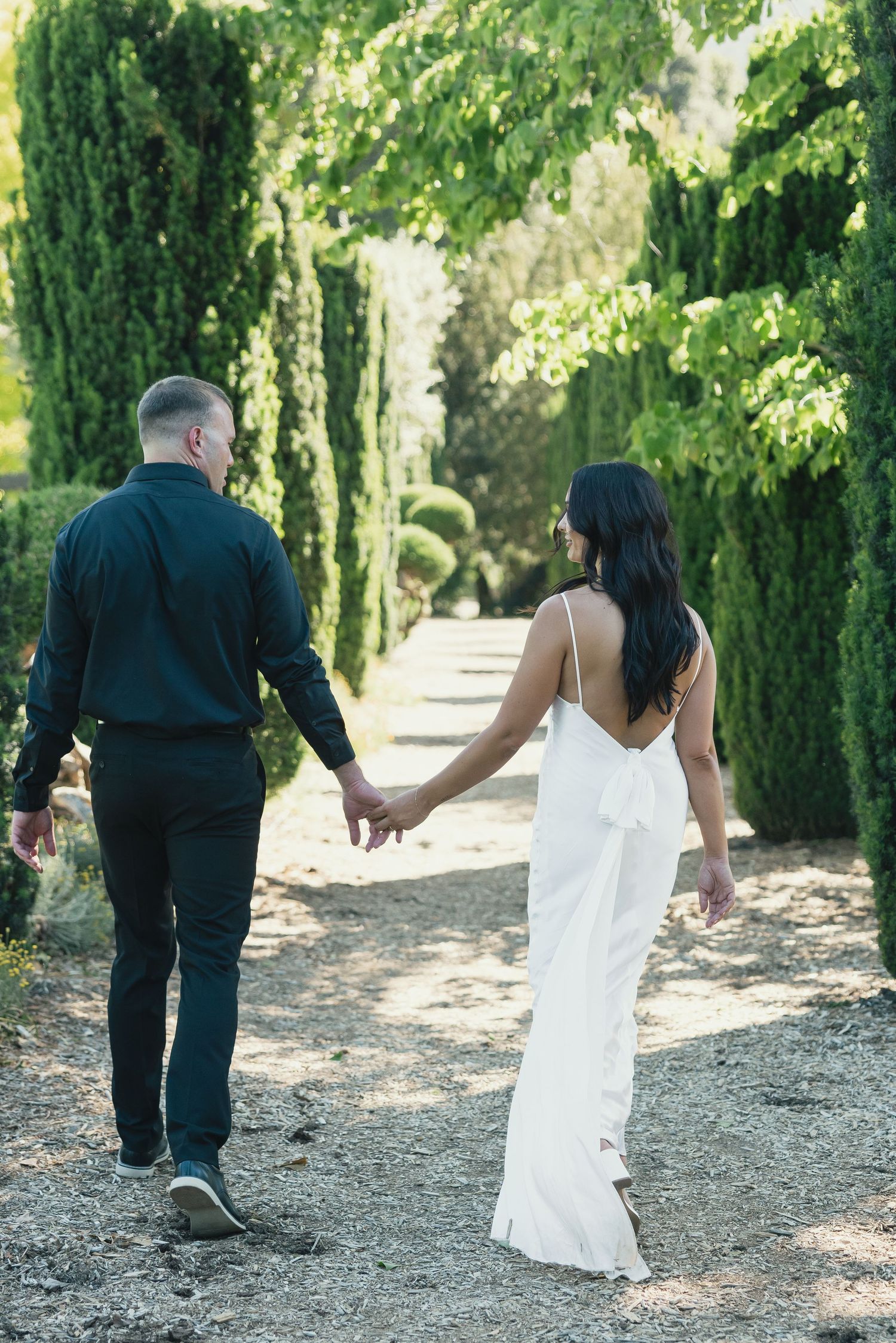 A couple walks along a cypress-lined path in formal wedding attire.