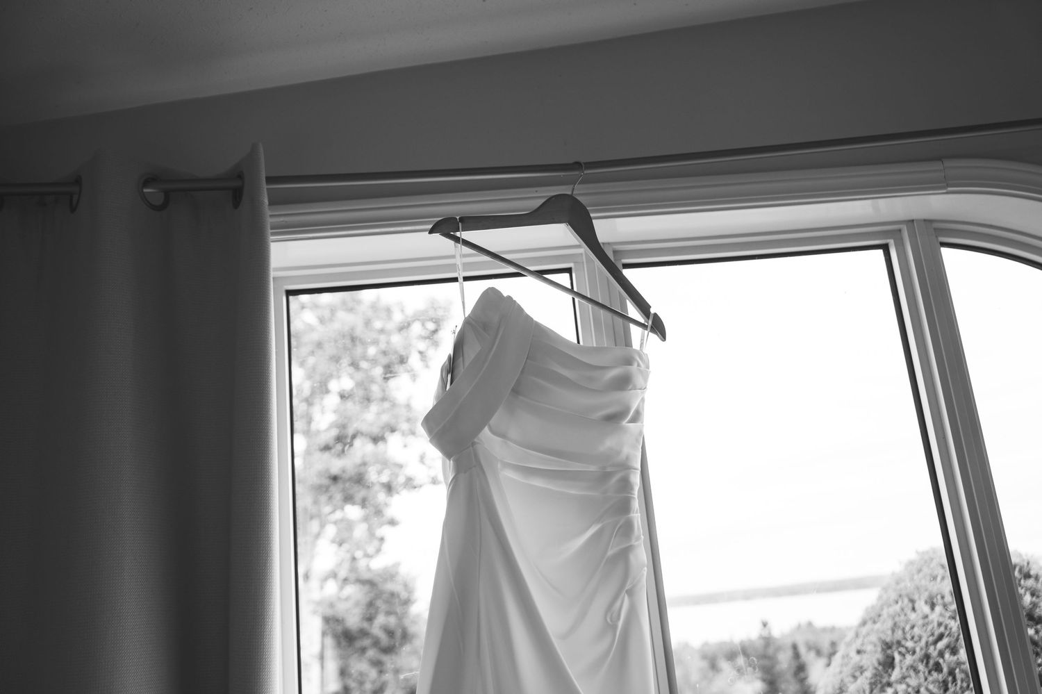 Black and white photo showing bride in flowing white wedding dress standing by window in bridal preparation room.