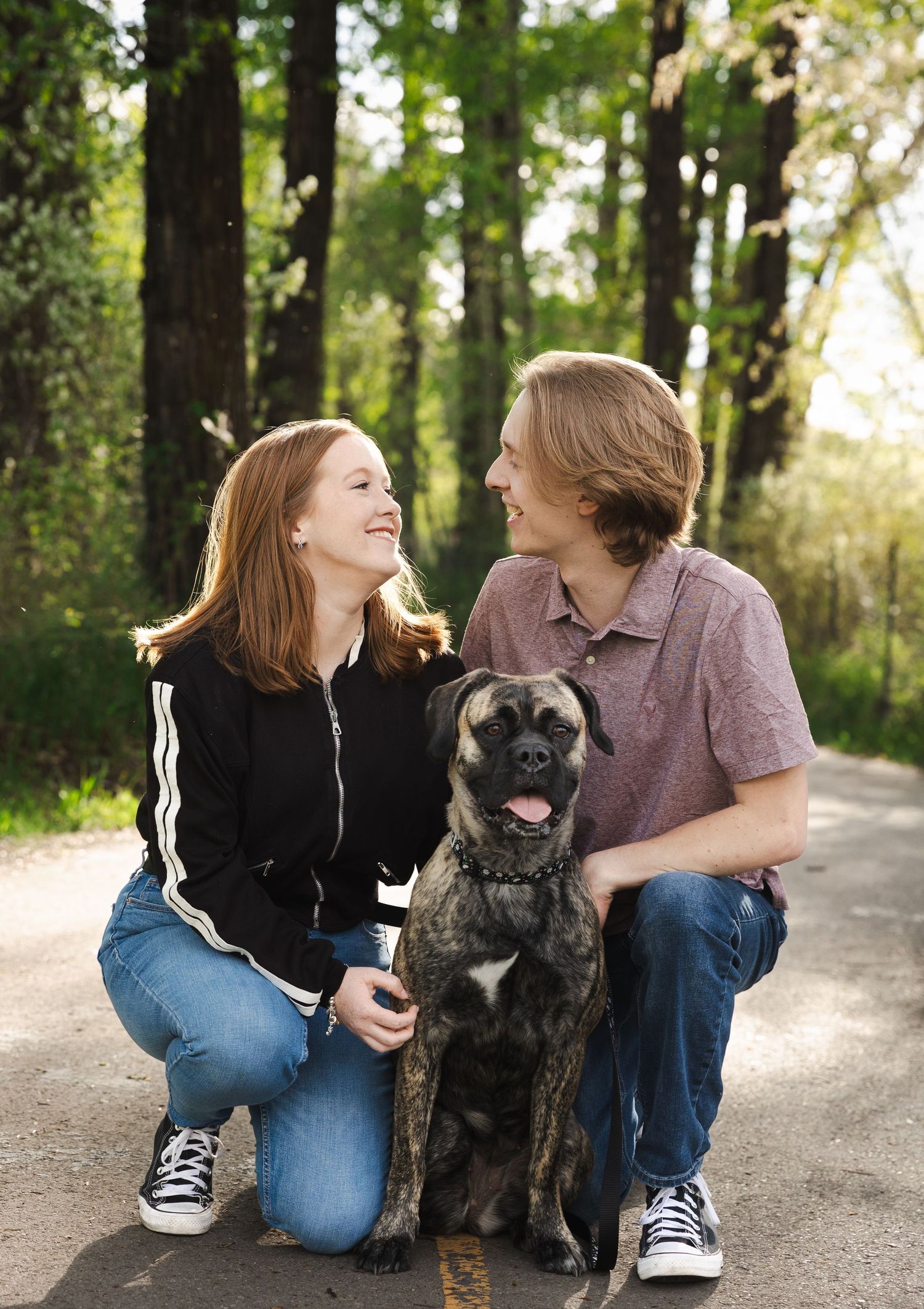 Couple sitting on a path with their black dog in a sunlit forest setting.