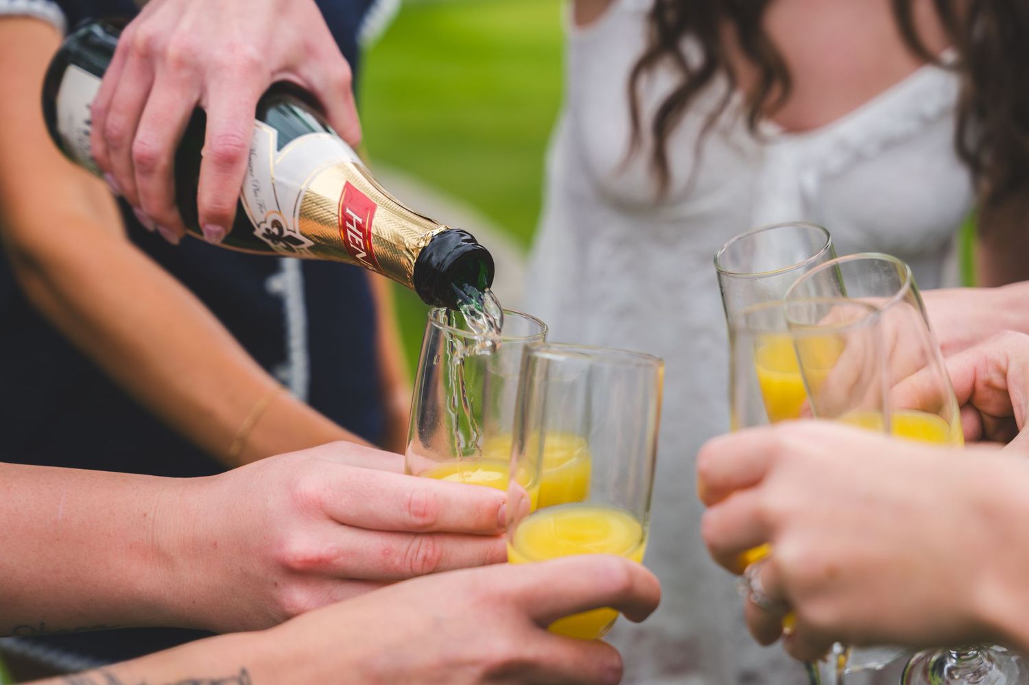Hands pouring drinks into glasses at a bar or party setting with multiple beverages being served.