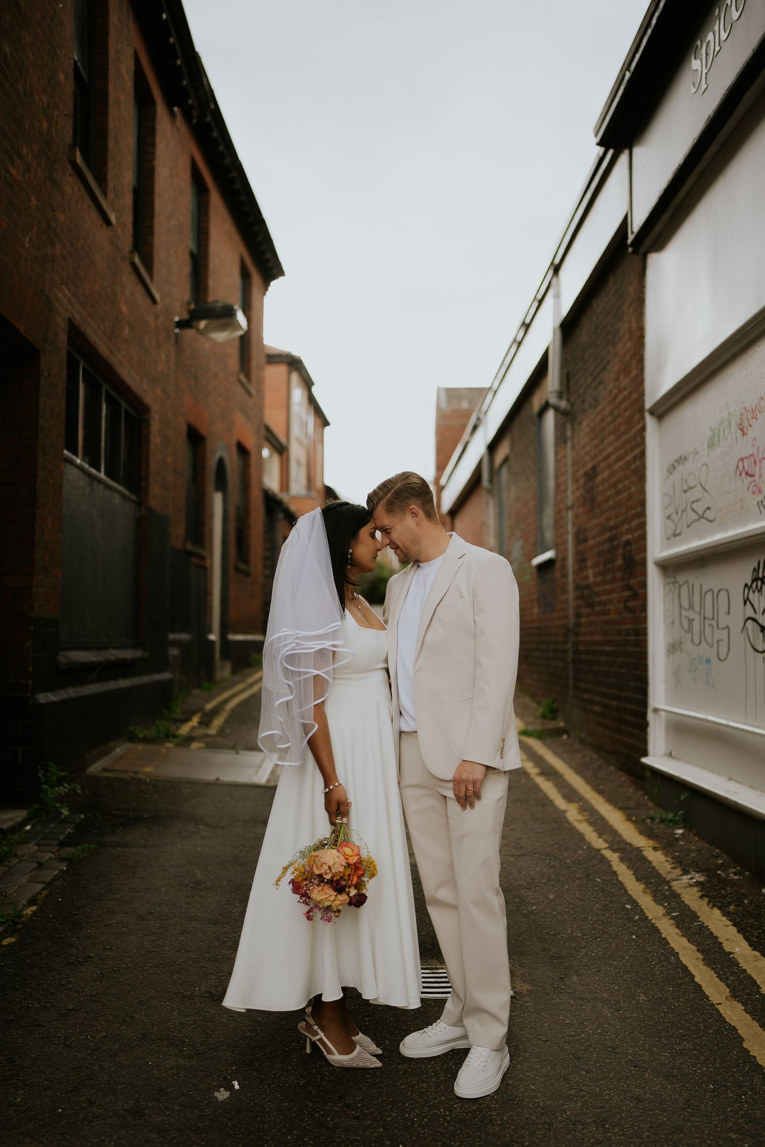 Bride in a white dress and veil holding a colorful bouquet touches foreheads with her groom in a cream suit in a brick alleyway.