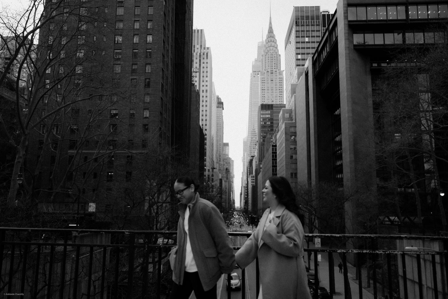 A romantic couple walks hand in hand across a bridge with the New York City skyline in the background, captured in black and white.