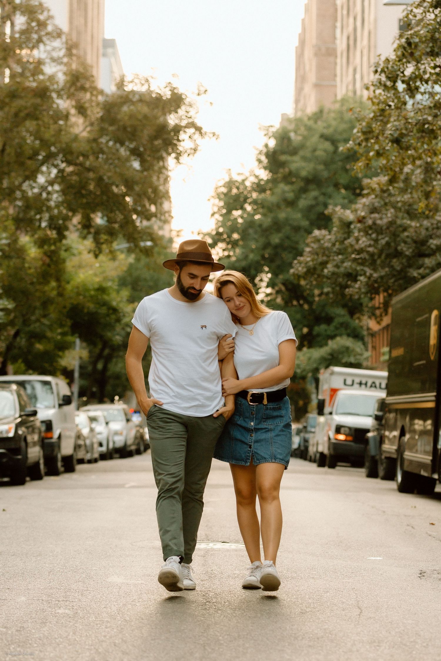 A couple walks casually down a New York City street during the day, dressed in relaxed outfits and sharing a candid moment.