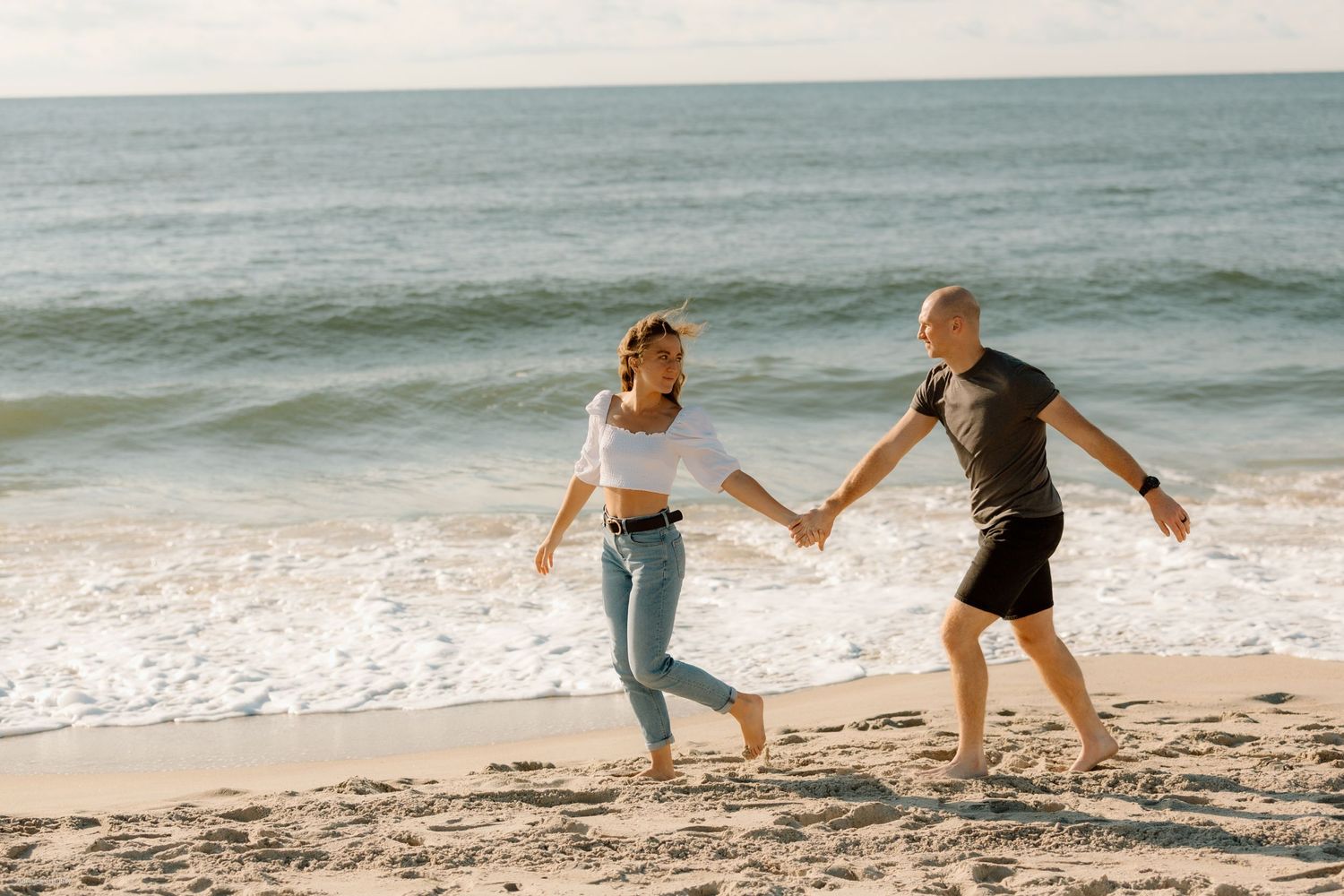 A joyful couple runs hand in hand along the destination beach, laughing together as waves roll in behind them.