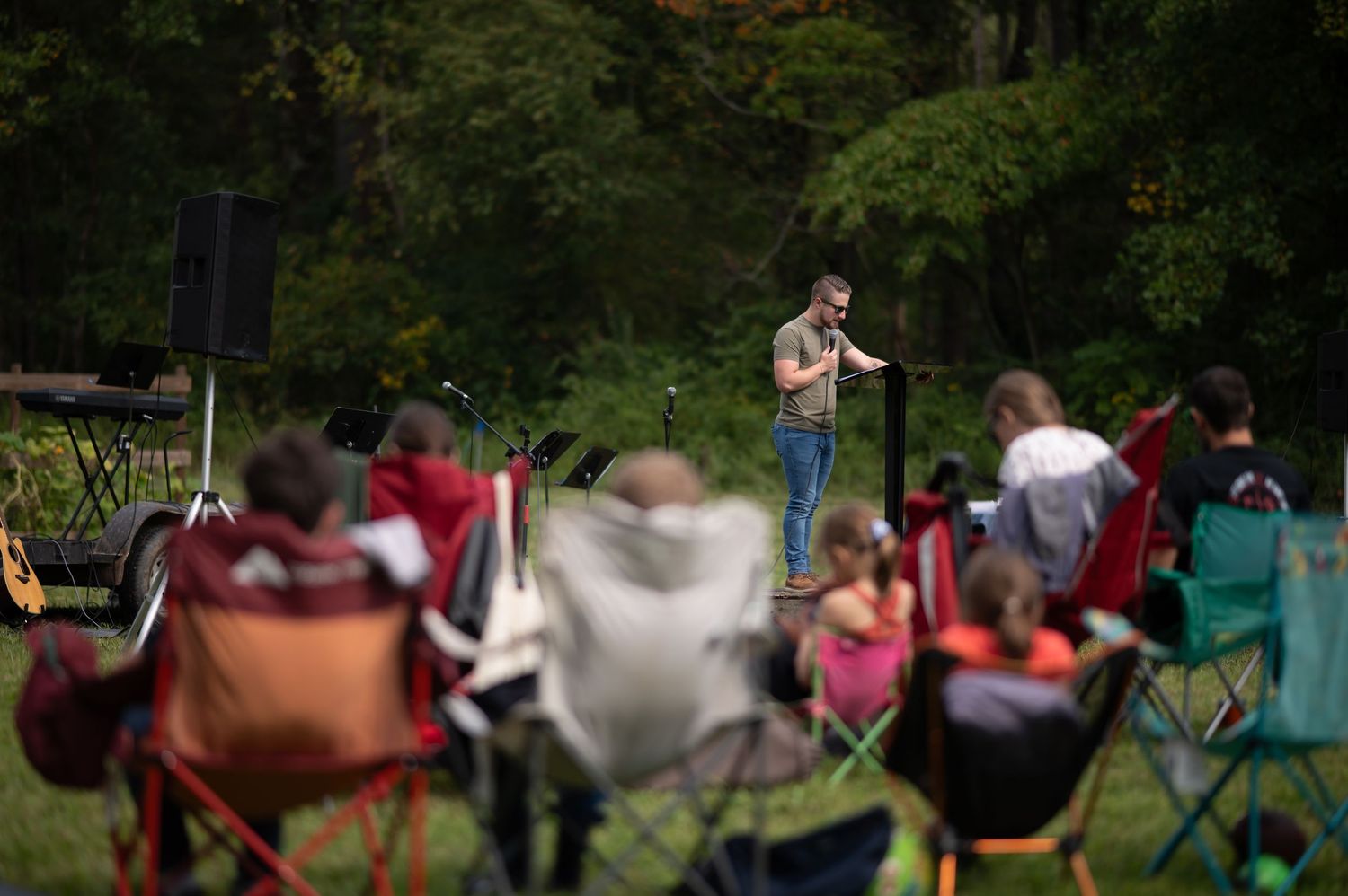 Pastor Brandon preaches to a crowd of Crossroads members sitting in camp chairs at church on the trail, outside in a green field