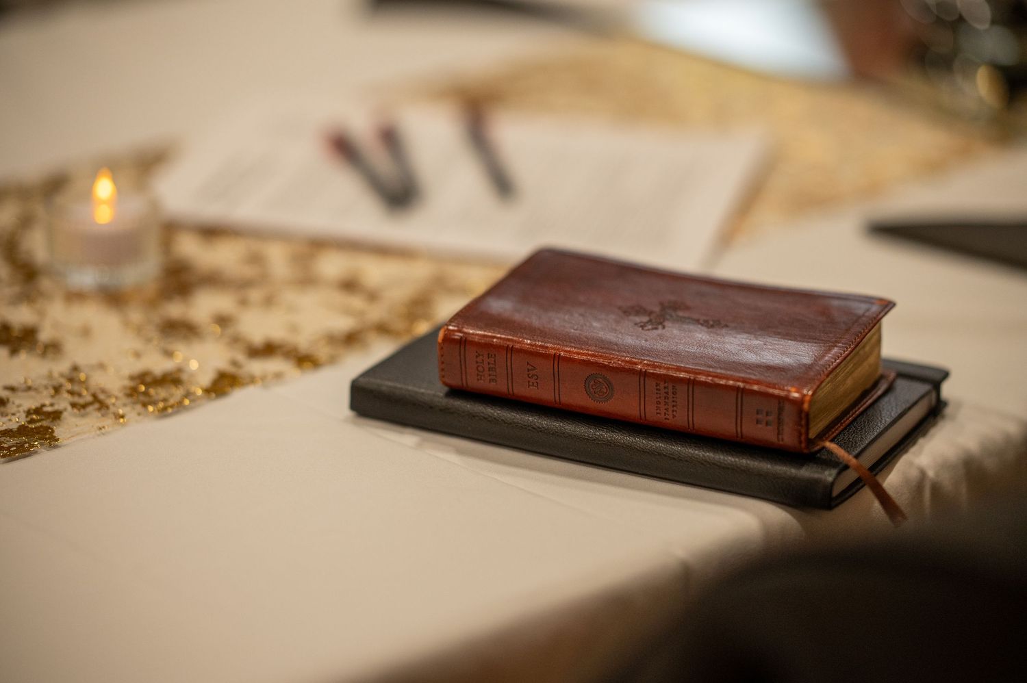a small brown leather esv version Bible and a compact black leather notebook lie on a white tablecloth with a gold candle in the background