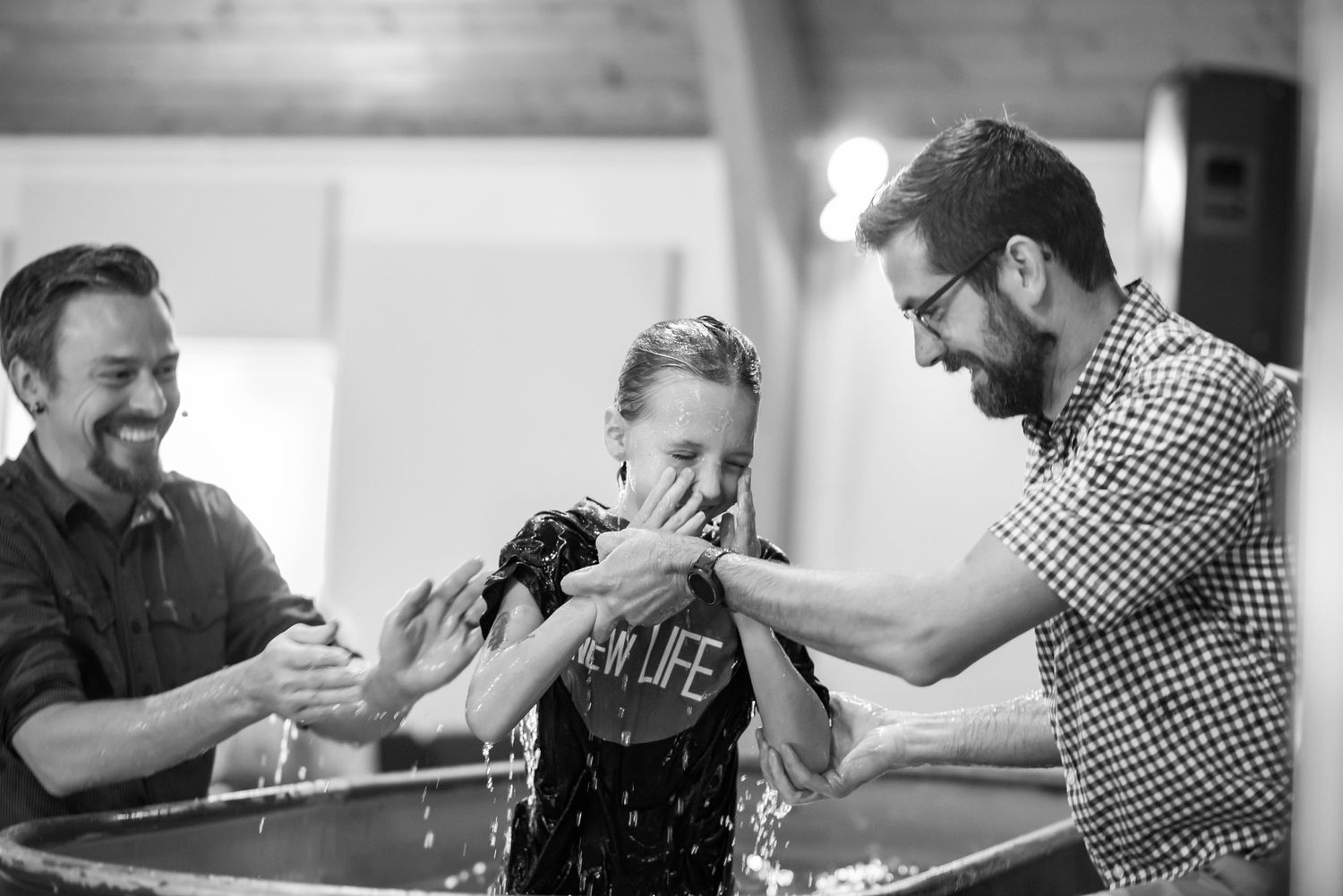 black and white image of a baptism of a young girl at Crossroads church; she rises from the water as two pastors help her, smiling