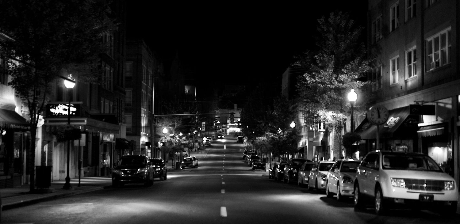 black and white image of High Street in downtown Morgantown at night, with streetlights shining along the street and parked cars