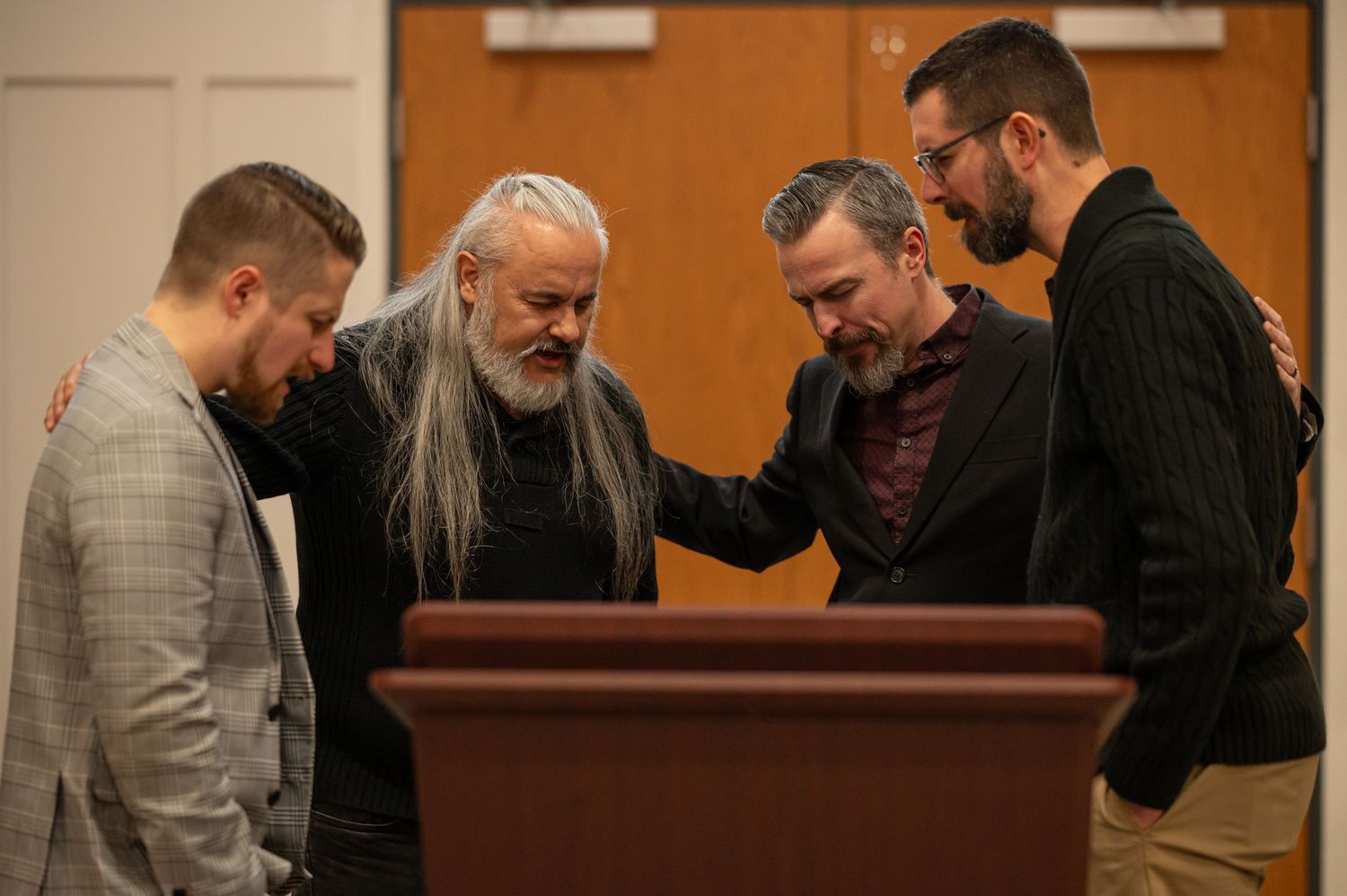 All four Crossroads pastors stand together behind a wood pulpit with arms around each others' shoulders, praying before Calibrate 2026