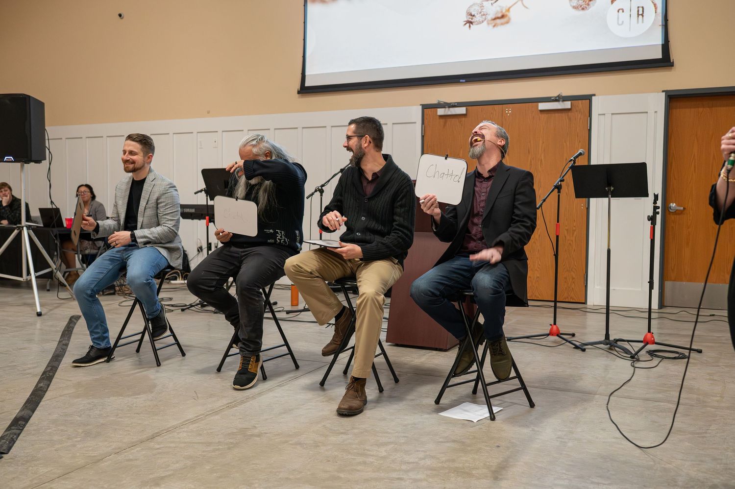 All four Crossroads pastors sit on stools in an event center, holding up small white boards and laughing as they play a game at Calibrate