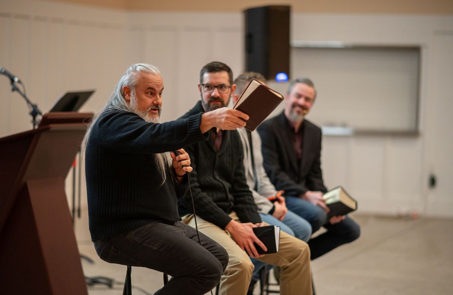 All four Crossroads pastors sit on stools as Pastor Chatto speaks into a mic while extending a Bible in his right hand.