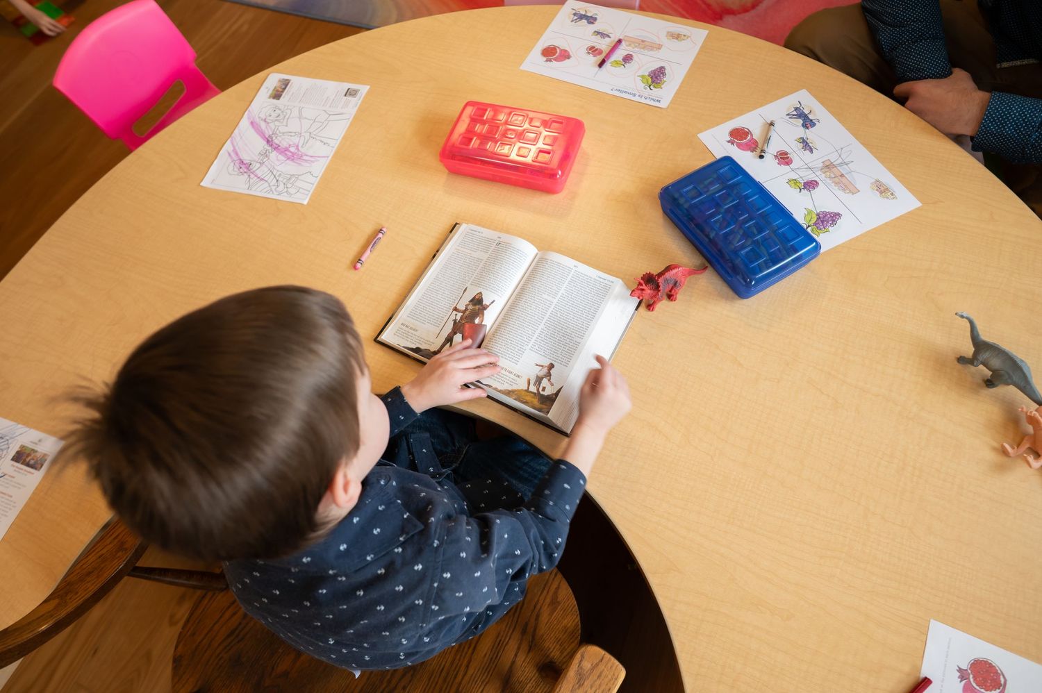 view from above of a little boy sitting at a large table, reading from an open Bible with crayons and coloring sheets scattered around