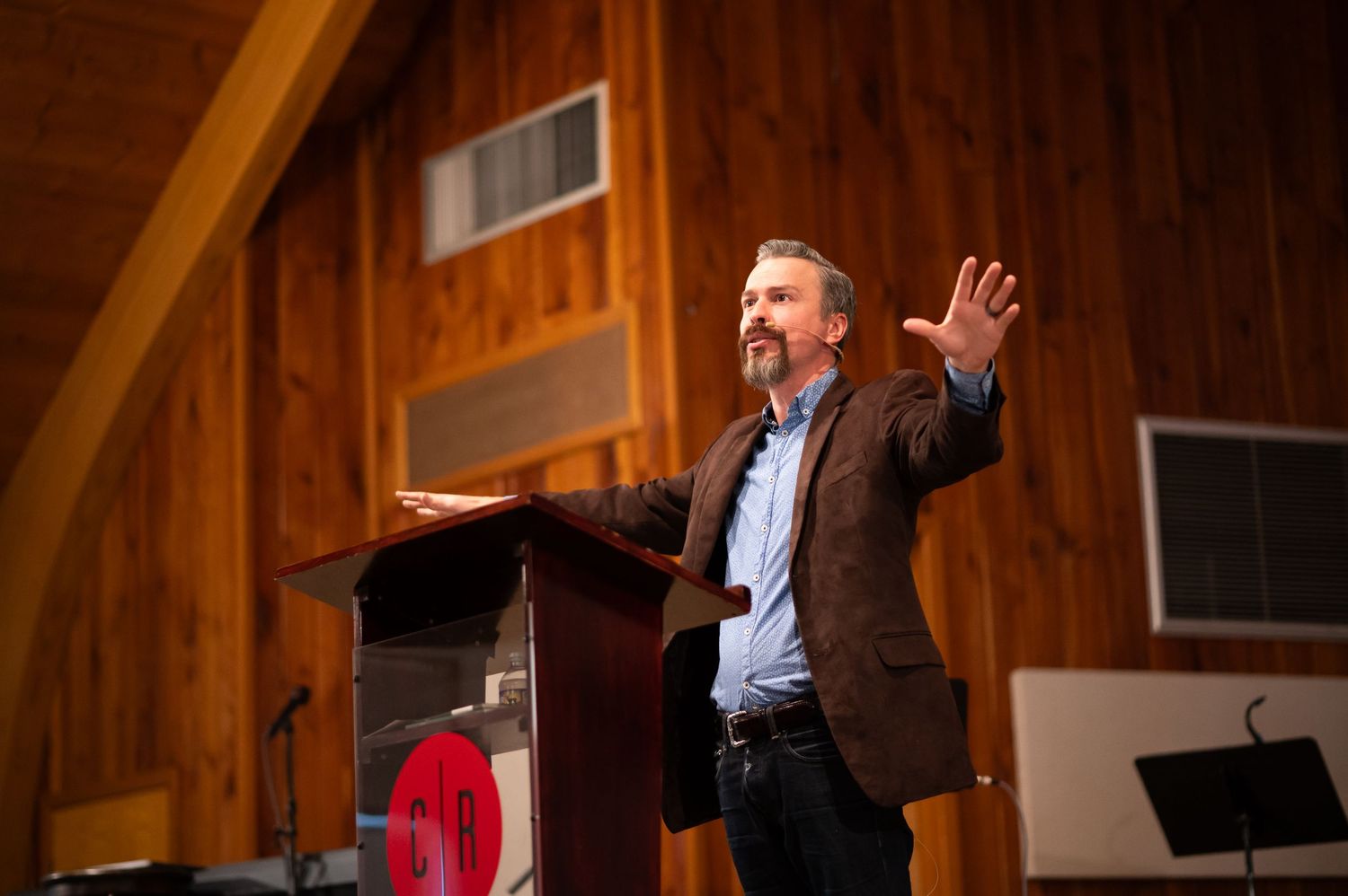 Pastor Chris preaches on the stage at Crossroads Morgantown with hands spread wide, and an open Bible on the pulpit in front of Him.