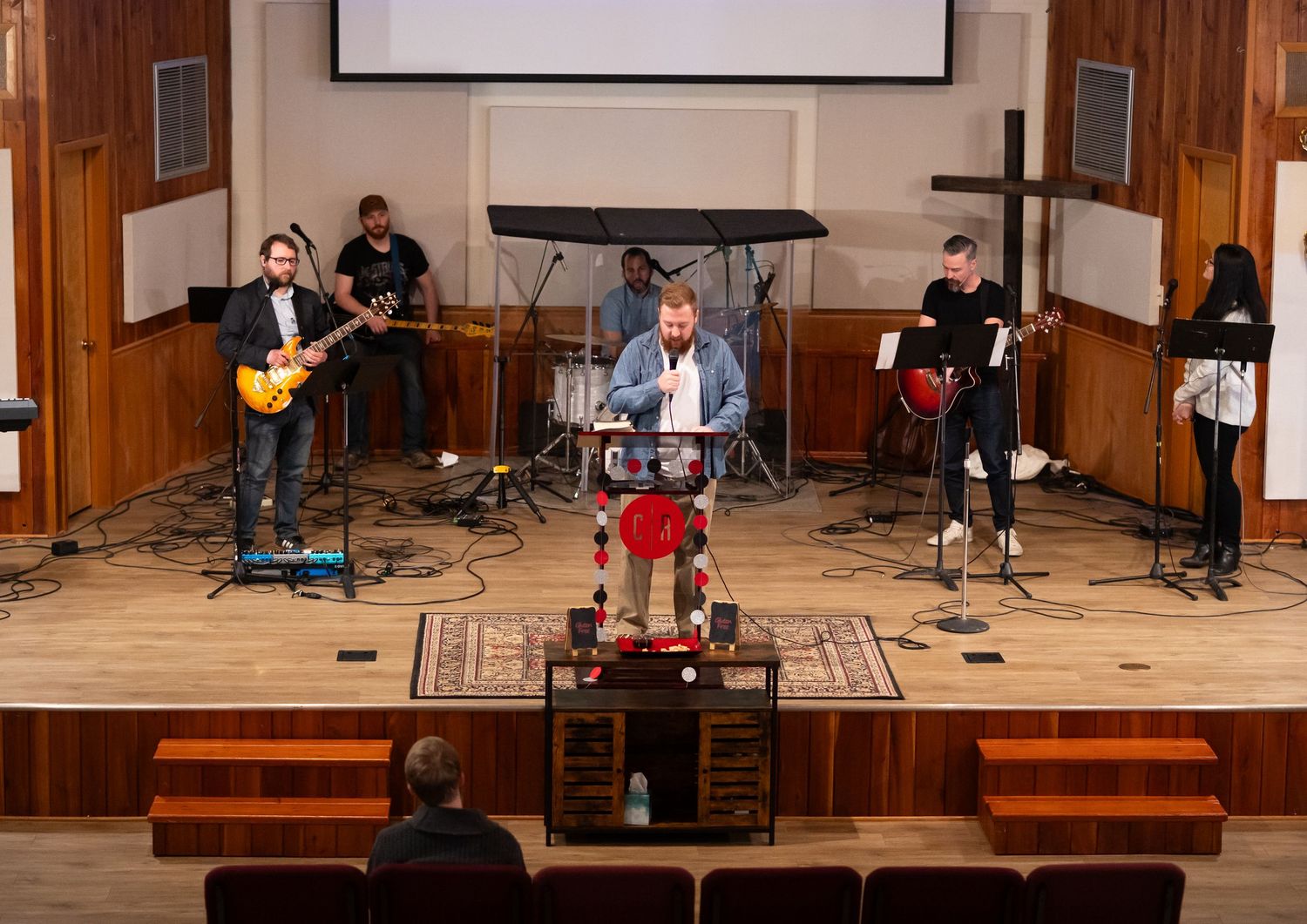 a church member stands at the pulpit and speaks at Crossroads Morgantown as the worship team stands ready with their instruments behind him