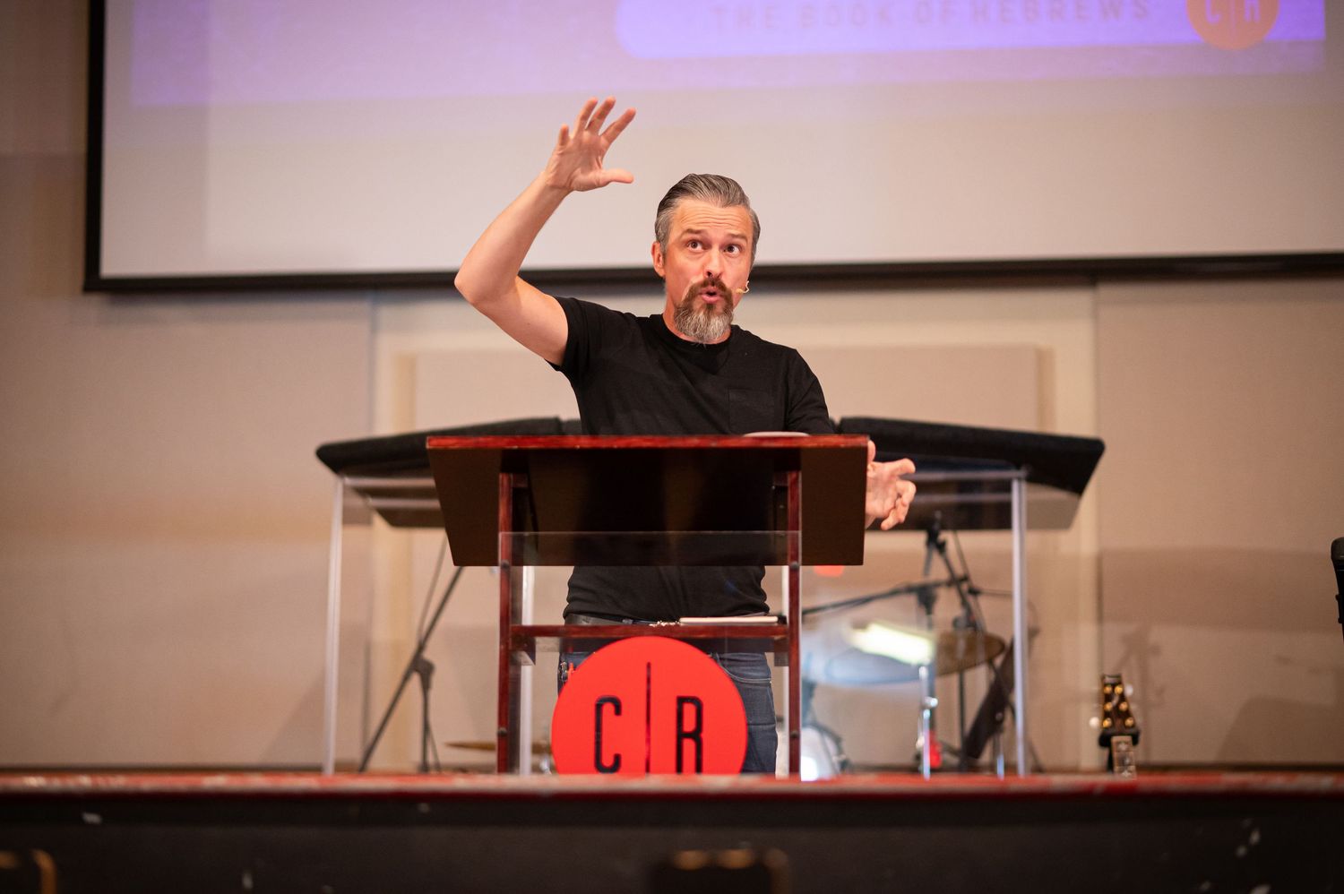 Pastor Chris preaches on stage at Crossroads Morgantown with one hand raised and the Crossroads logo visible on the pulpit in the foreground