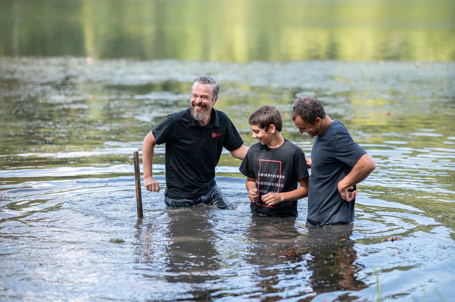 Pastor Chris stands in the river with a young church member and his father for a baptism at an outdoor Sunday morning service in Morgantown