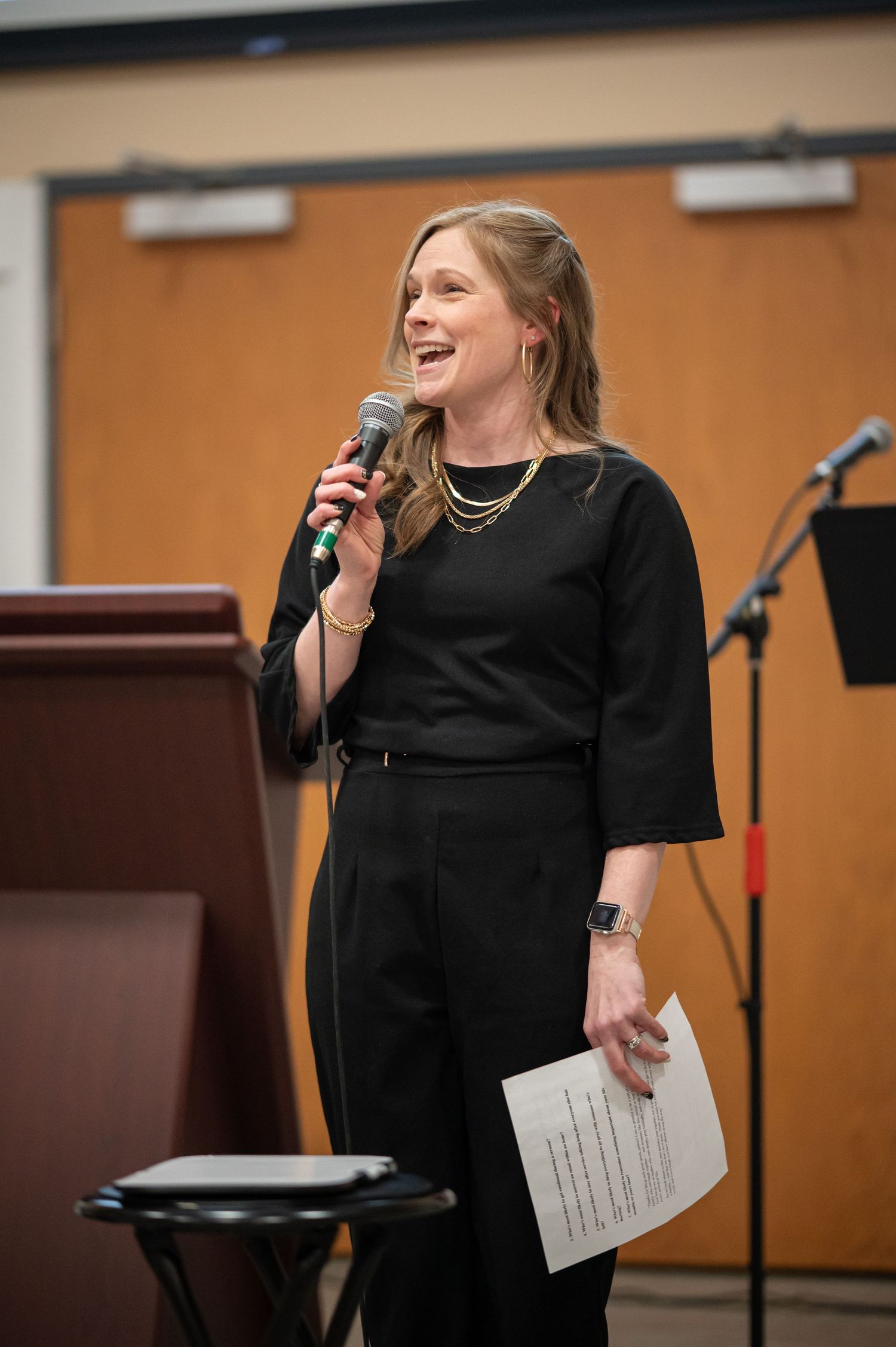 a woman in a black dress with long hair speaks into a microphone and laughs at a Crossroads member event