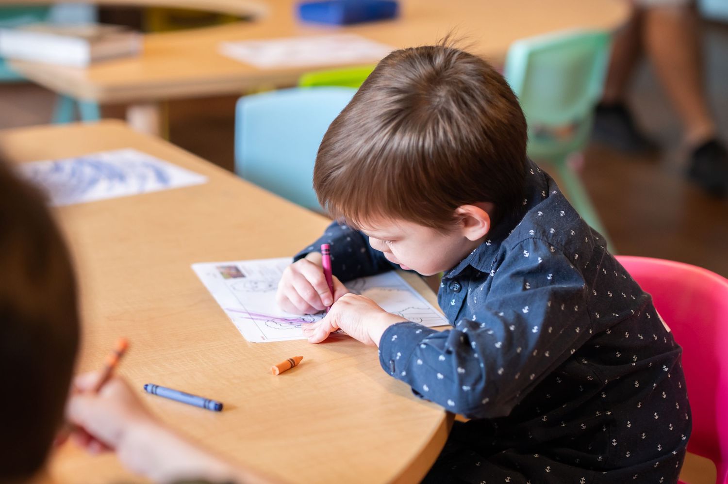 a little boy sits at a wooden classroom table and leans over a coloring sheet, coloring intently during kids' ministry at Crossroads Church