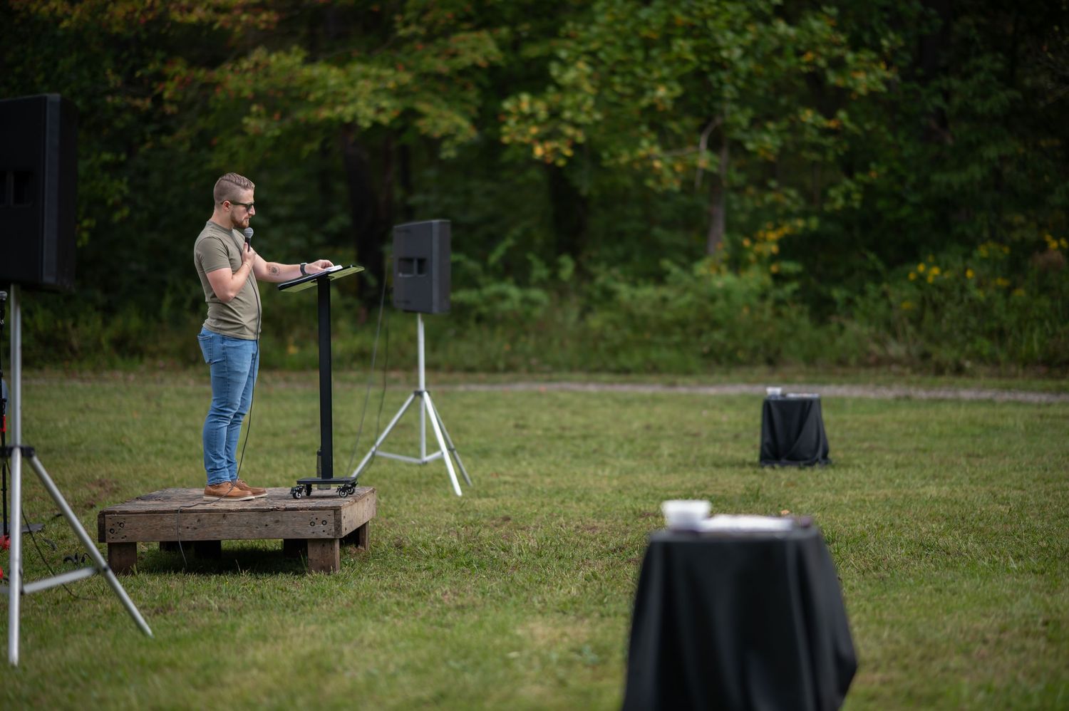Pastor Brandon stands on a small stage in a large field, preaching during an outdoor church service at Crossroads.