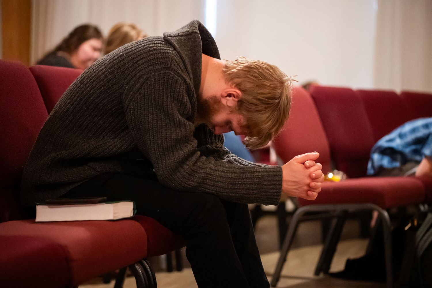 a young man seated in red church chairs bows his head and prays with hands clasped, during Sunday morning church service in Morgantown, WV