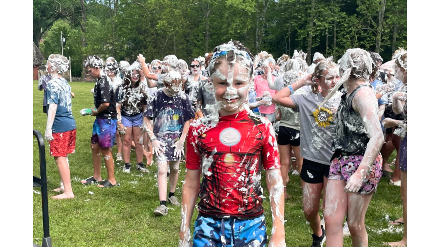 a boy covered in shaving cream stands in the grass and smiles for the camera, with many other kids covered in shaving cream behind him