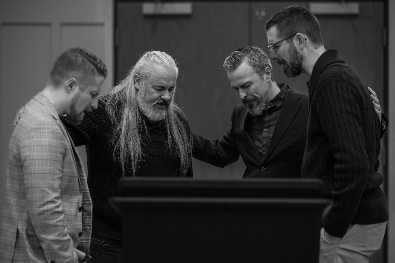 Black and white candid photo of all four pastors praying together around a pulpit, with heads bowed and eyes closed.