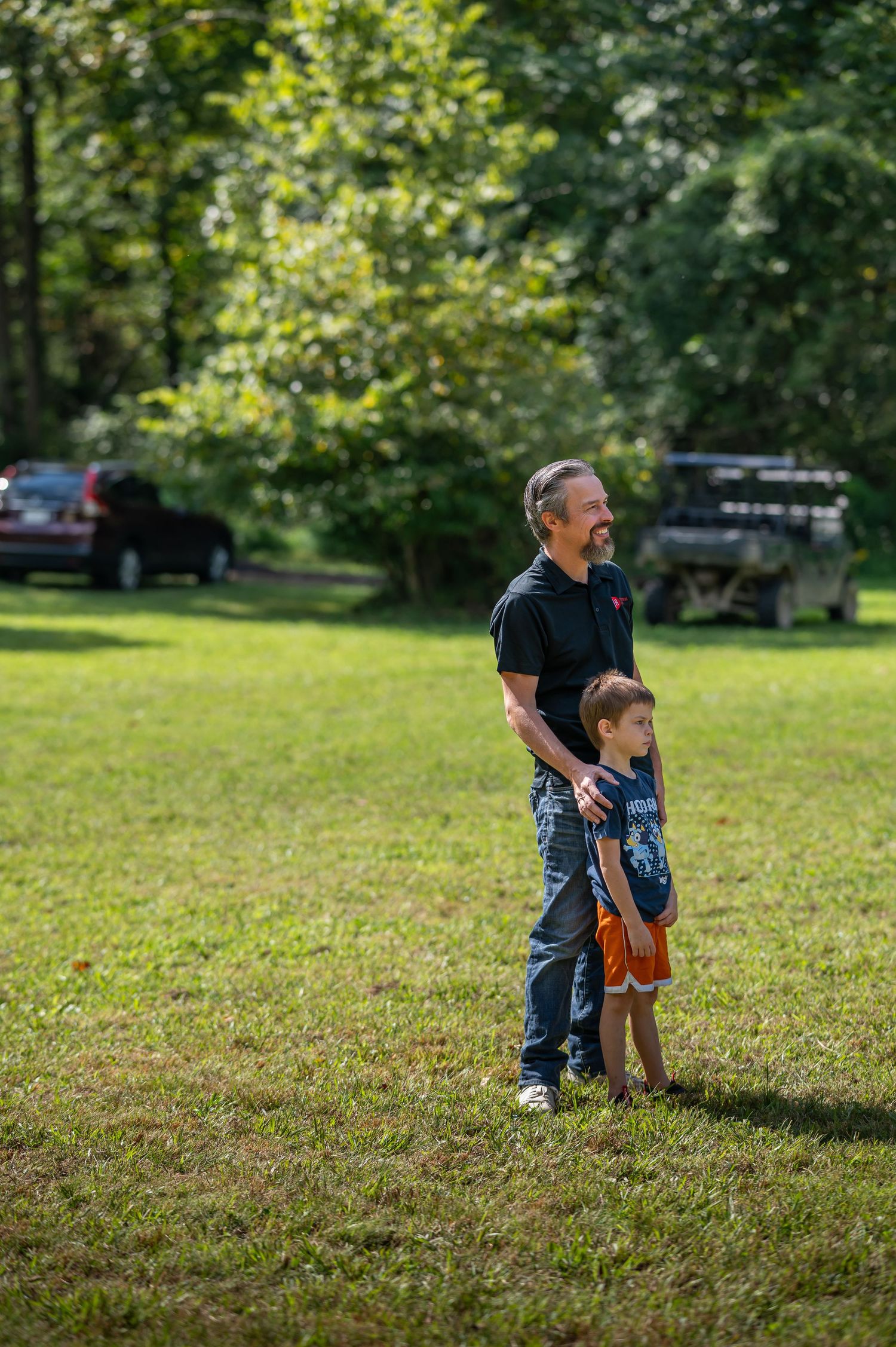 Pastor Chris stands outside in the grass with his little son, watching another pastor preach during an outdoor Crossroads church service