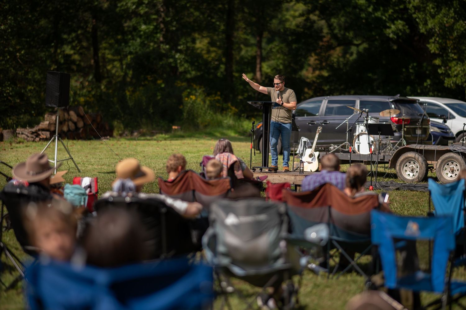 Pastor Brandon preaches from a small stage during an outdoor church service with church members in camp chairs sitting listening to him