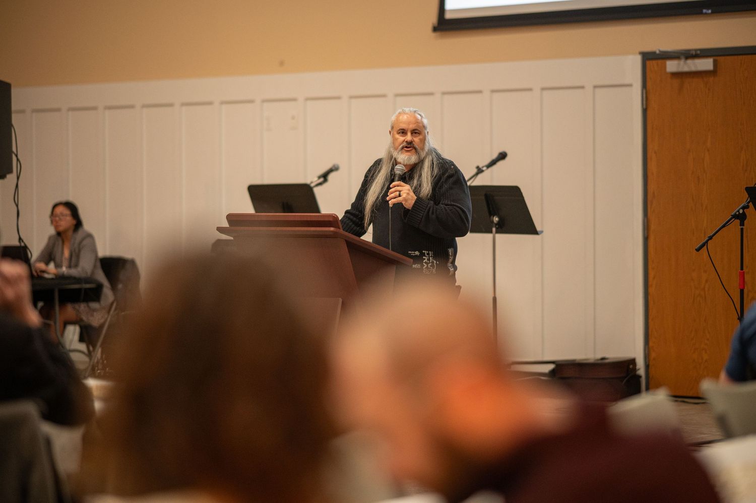 Pastor Chatto stands behind a wooden pulpit at an event center with microphone in hand
