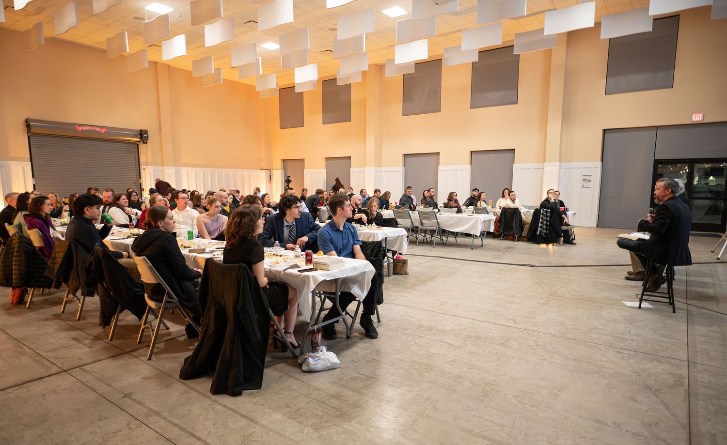 Church members sit in an event hall at long tables covered in white tablecloths, as Crossroads pastors speak from the front of the room