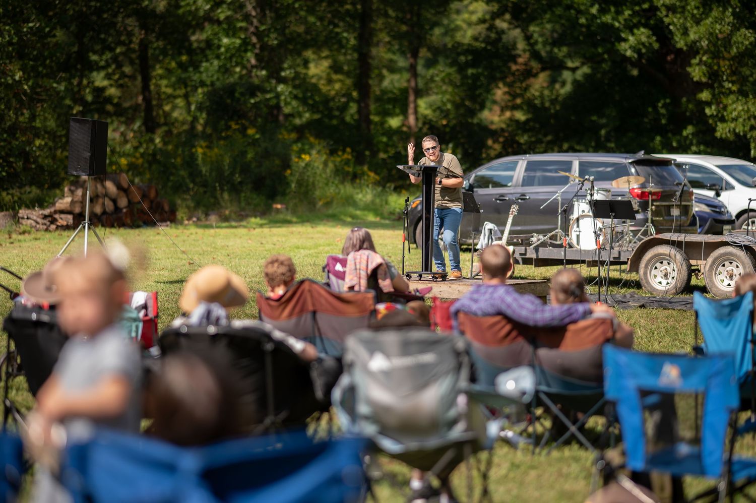 Pastor Brandon preaches from a small stage in an open field during an outdoor church service as church members sit in camp chairs and listen