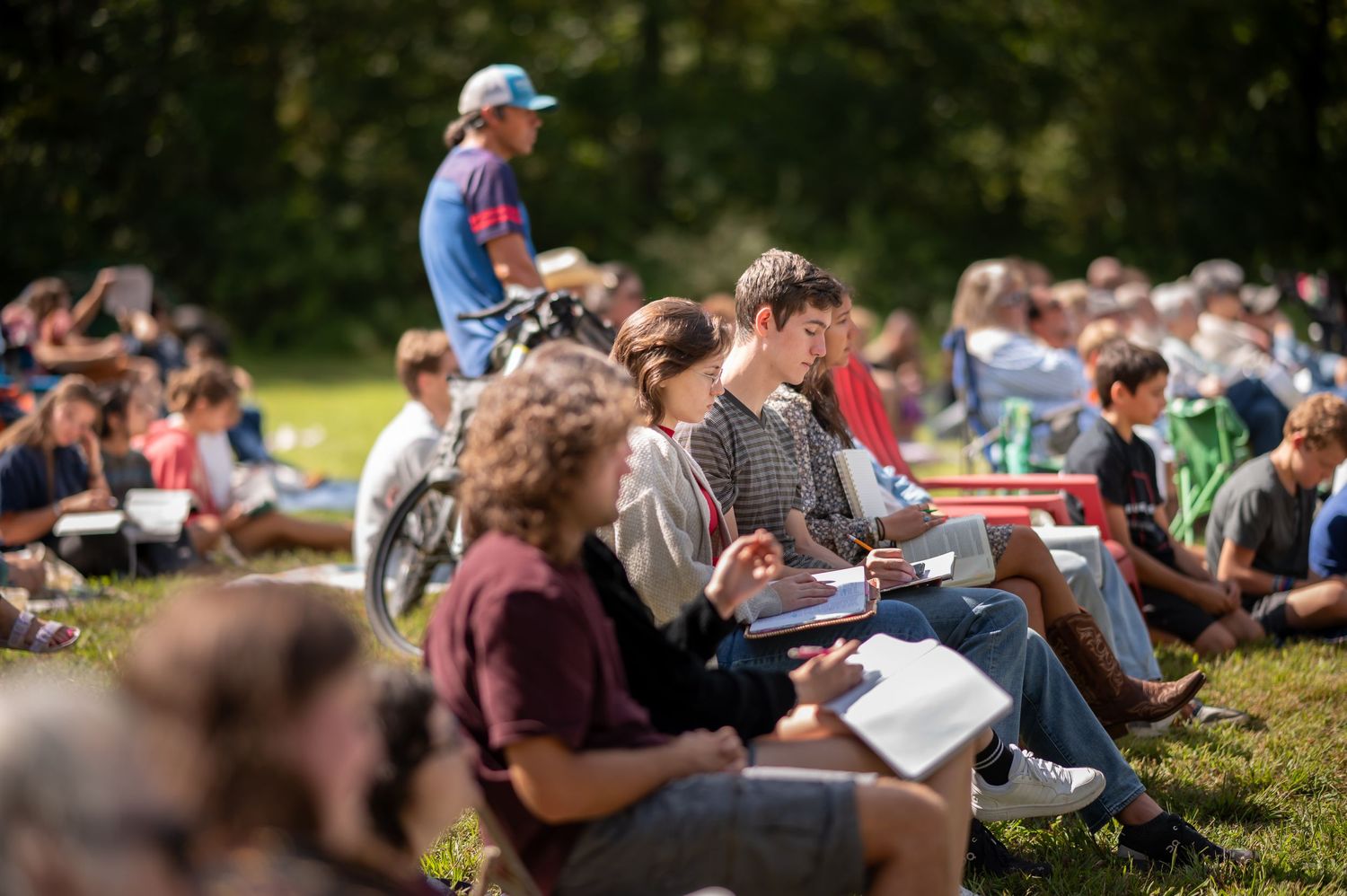 church attendees stand and sit in the grass listening to preaching during an outdoor Crossroads church service in Morgantown, WV