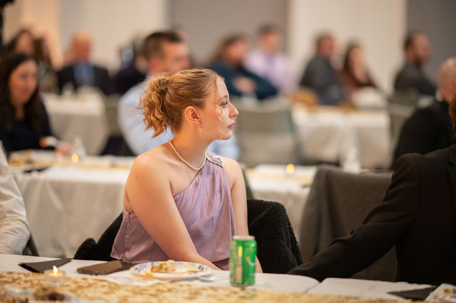 a young woman sits at a table in a conference center, listening intently to the speaker on stage