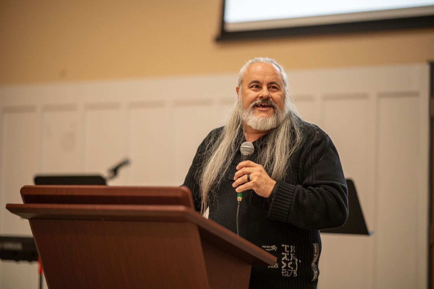 Pastor Chatto stands at a pulpit in an event center, speaking at the Crossroads annual members' meeting