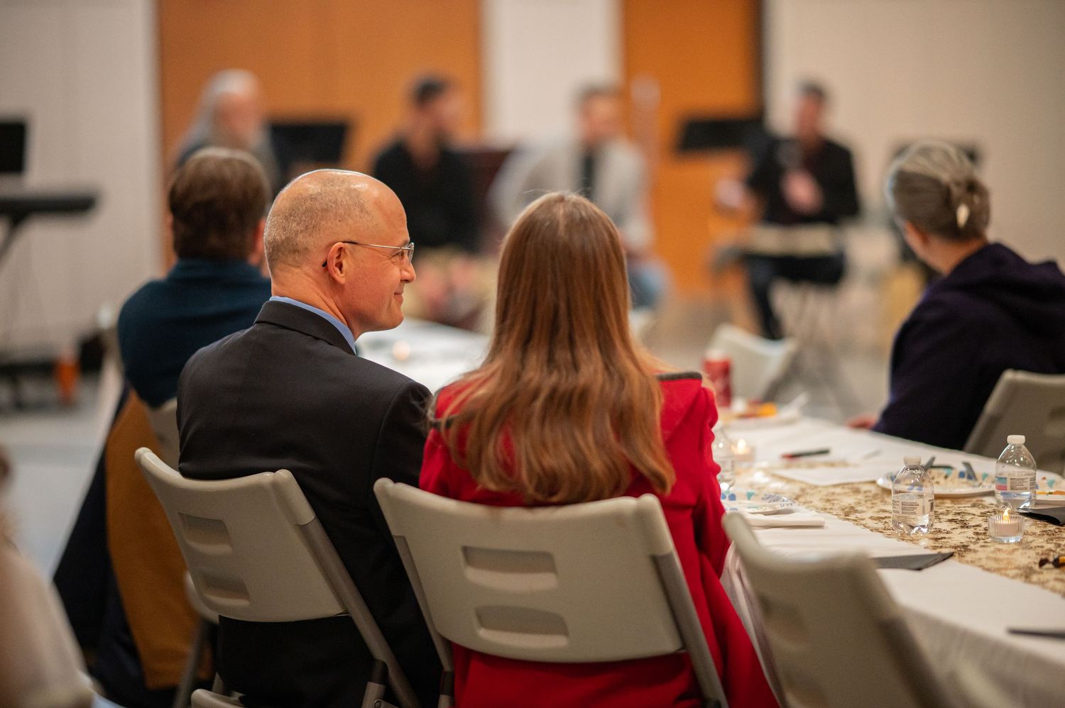 a husband and wife sit together in folding chairs at a Crossroads event, listening to the speaker