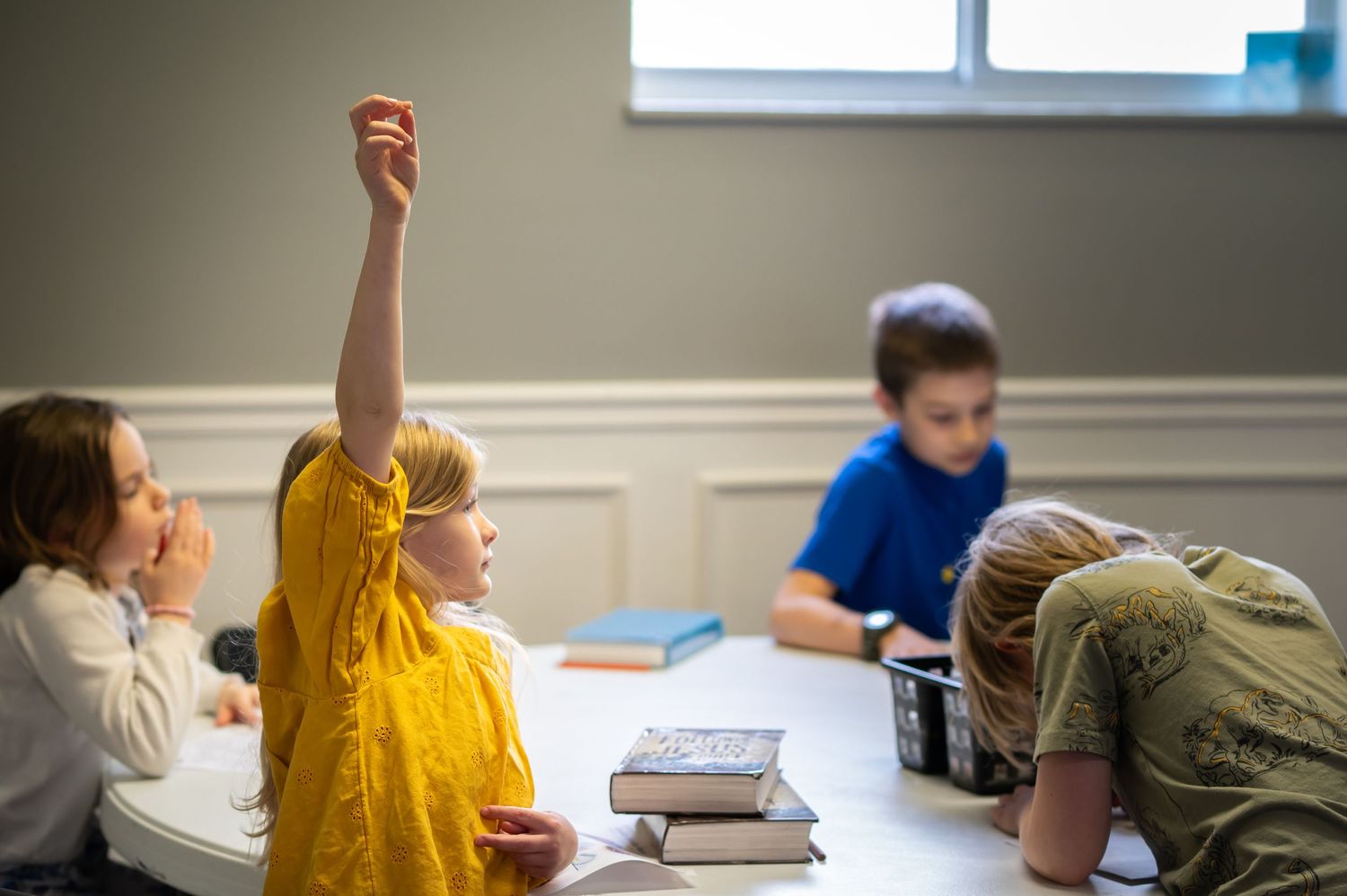 a little blonde girl raises her hand high to answer a question as she sits at a table during kids' ministry during Sunday church service