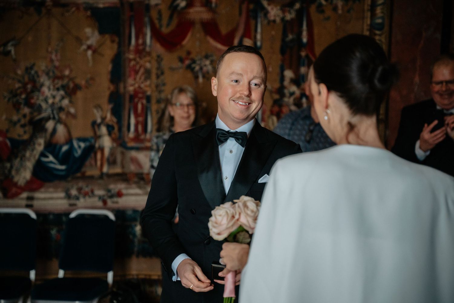 Two people in formal attire converse during an elegant evening event with ornate decorations in the background.