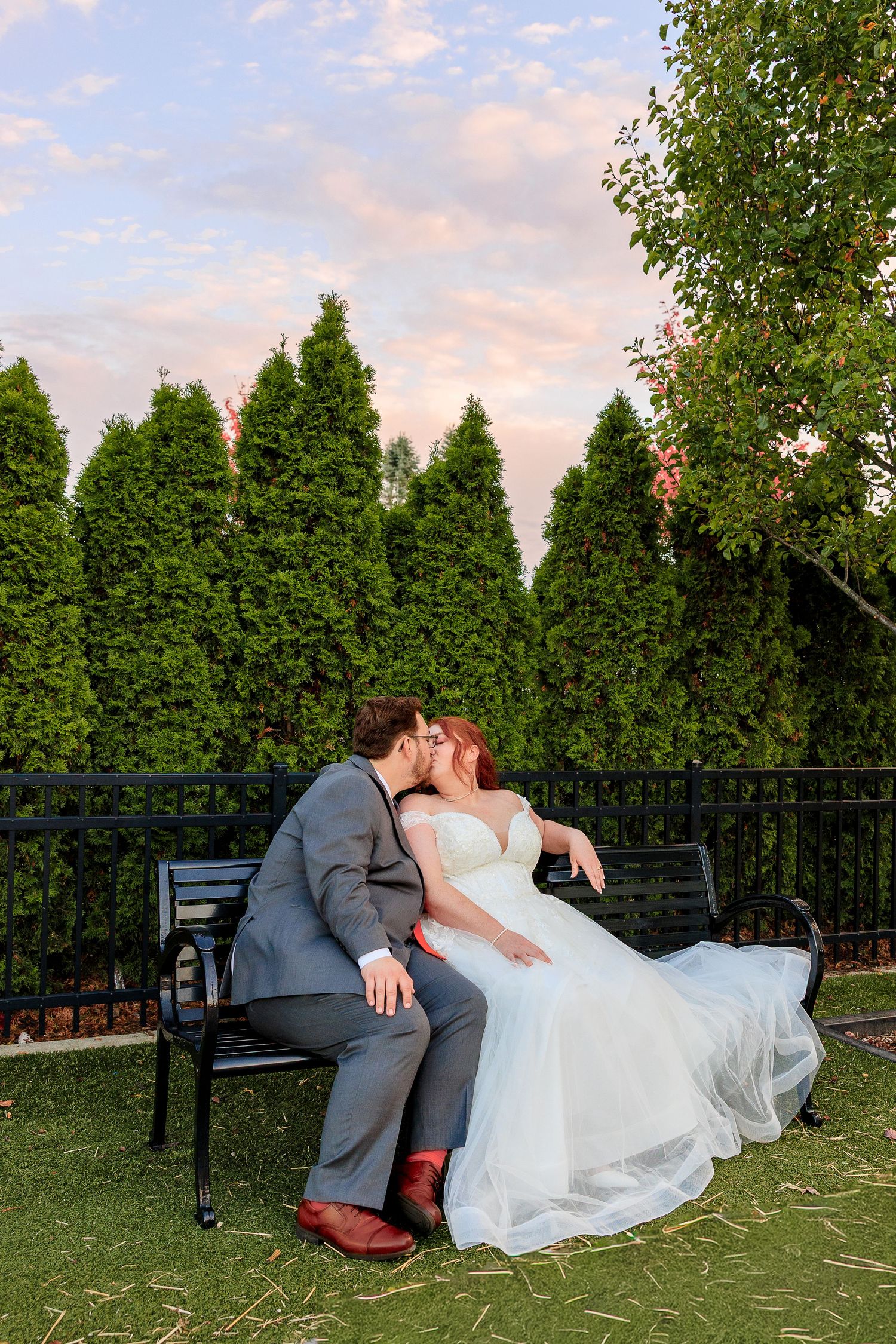 A romantic wedding couple shares a tender dip pose on a lawn with green trees in the background at sunset.