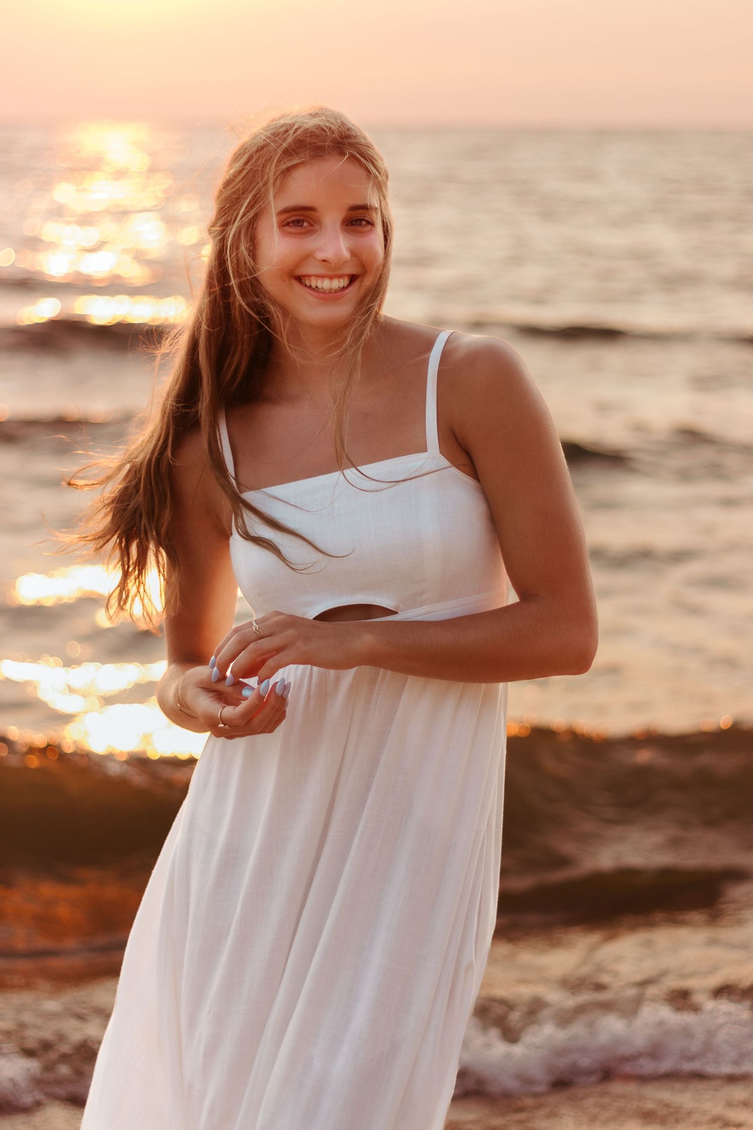 Woman wearing white dress smiling on beach during golden sunset.
