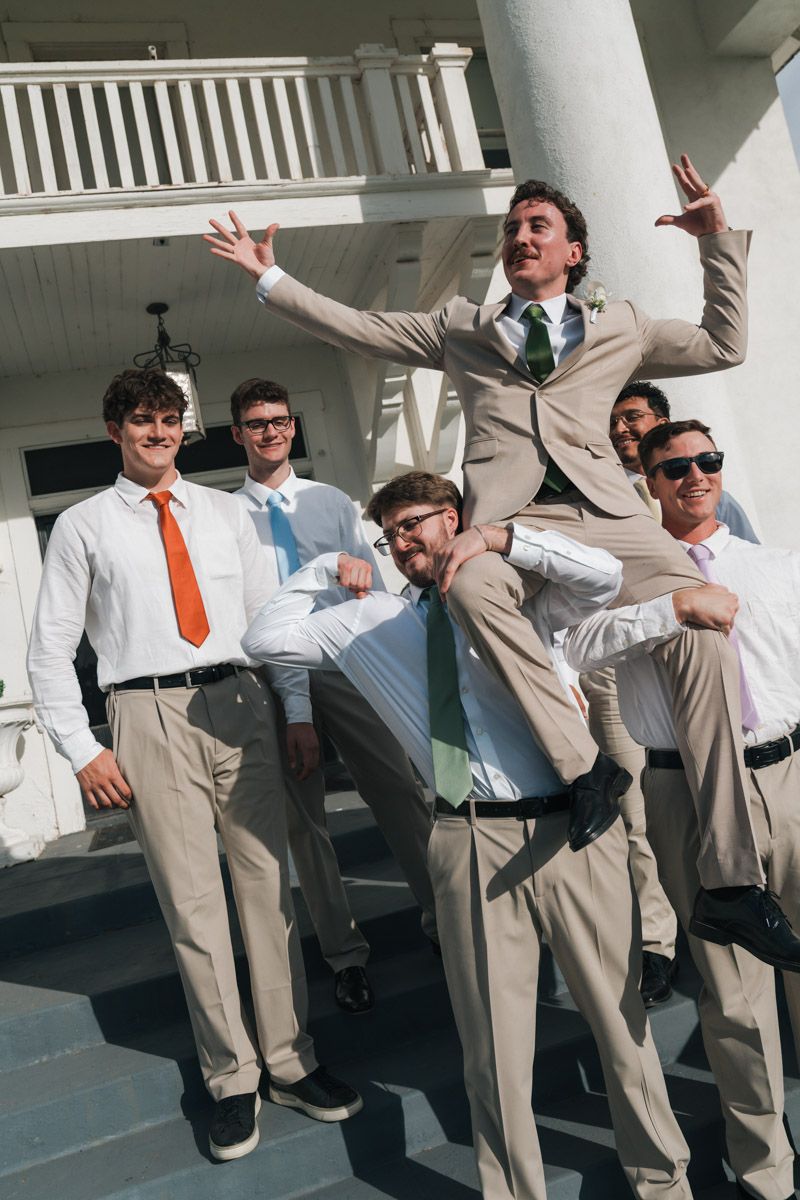 Group of young men in suits posing playfully outside a white building, one being lifted by friends.