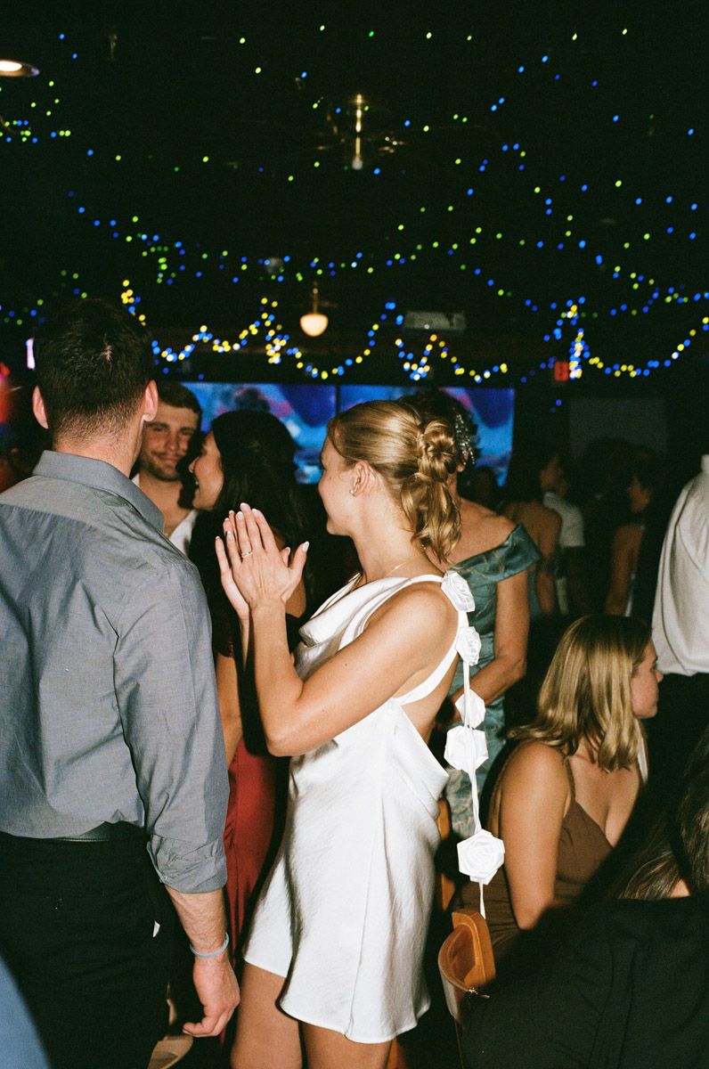 Woman in white dress with floral detail dances at elegant evening party with colorful string lights.