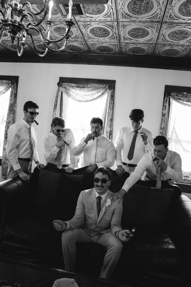 Groom and groomsmen in white shirts and sunglasses with cigars in an elegant room before wedding ceremony.