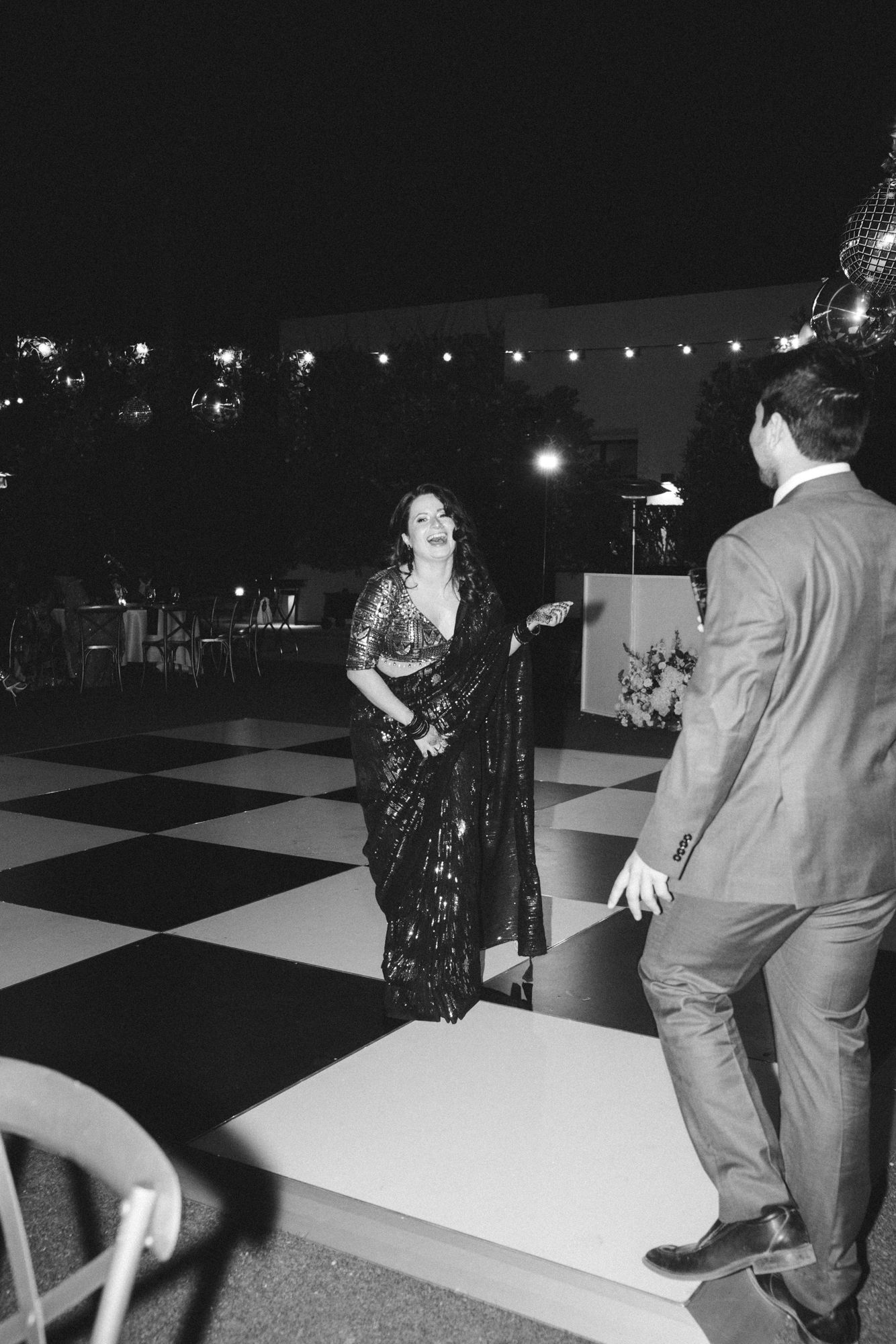 Woman in sequined saree dancing with suited man on checkered dance floor at elegant evening reception.